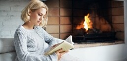 Une femme lit un livre près d’un foyer.

