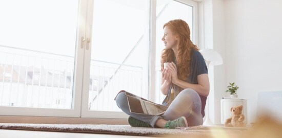 A woman sitting on a rug, holding a tea cup and looking out glass patio doors.
