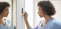 Une femme ajuste un thermostat à côté d’un miroir.
