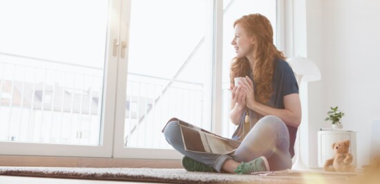 A woman sitting on a rug with a magazine and a teacup, looking outside glass patio doors.
