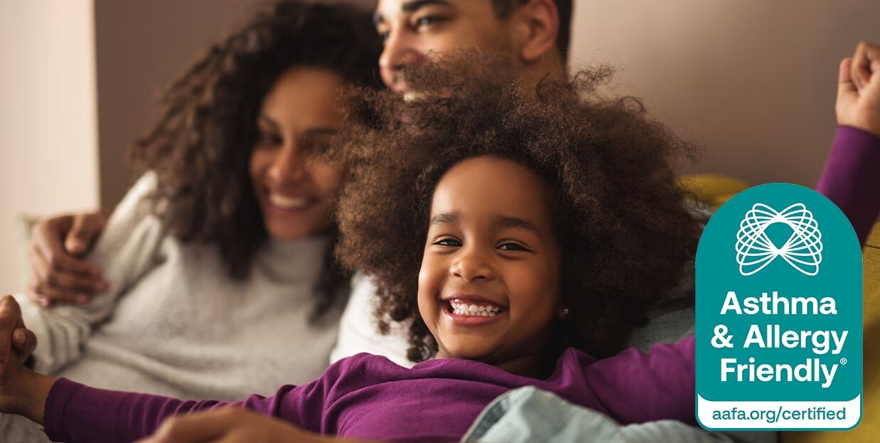 A family sitting close together on a couch with blurred faces, accompanied by an Asthma & Allergy Friendly Certified seal on the right side of the image.
