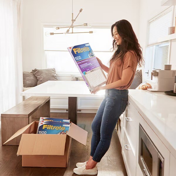 Une femme debout dans une cuisine lumineuse tenant un filtre à air Filtrete, plusieurs filtres supplémentaires étant placés dans une boîte ouverte sur le sol.
