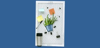 A furnace filter with various office supplies, a plant, and a travel mug on it.

